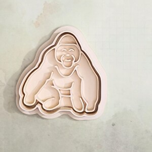 Könnte beinhalten: Hellbeige Gorilla-Ausstecher. Der Ausstecher hat ein mehrschichtiges Design, wobei der Gorilla in sitzender Position lächelt. Der Ausstecher ist aus Kunststoff und eignet sich zur Herstellung von Keksen oder anderen Leckereien.
