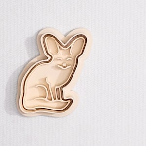 May include: A light beige cookie cutter shaped like a fennec fox. The cutter features a layered design with a dark brown outline and a smiling face. The fox is sitting with its large ears perked up.