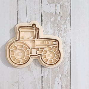 May include: A light beige tractor-shaped cookie cutter with a defined outline and internal structural details. The cutter features a large rear wheel, a smaller front wheel, a cabin area, and a hood section, all constructed with a raised edge.