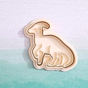 May include: A beige plastic cookie cutter shaped like a dinosaur. The cutter has a detailed design with multiple layers.