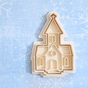 Könnte beinhalten: Beiger Kunststoff-Ausstecher in Form einer Kirche mit einem Kreuz oben. Der Ausstecher hat ein mehrschichtiges Design, bei dem der Umriss der Kirche und die Innendetails sichtbar sind. Der Hintergrund ist hellblau mit weißen Schneeflockenmustern.
