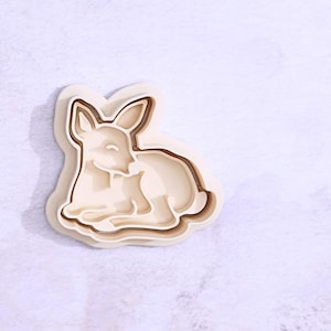 May include: A layered cookie cutter in the shape of a resting deer. The cutter is made of light beige plastic with a darker brown outline. The deer is lying down with its head turned to the side, and its eyes are closed.