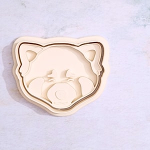 May include: A white plastic cookie cutter shaped like a red panda face. The cookie cutter has a detailed design with a cute expression.