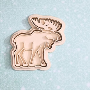 May include: A light brown moose-shaped cookie cutter. The cutter has a layered design, with the moose's antlers and body clearly defined. The background is a light blue with a subtle floral pattern.