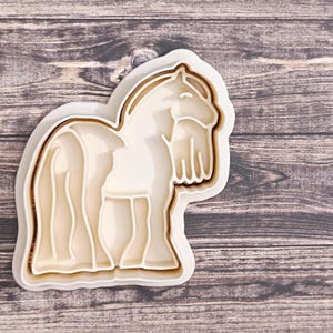 May include: A three-layer cookie cutter shaped like a horse. The horse is shown in profile with a long mane and tail. The cutter is made of white and tan plastic, ideal for baking horse-shaped biscuits.