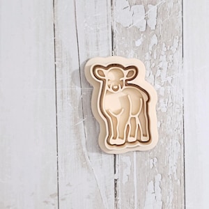 May include: A light beige cookie cutter shaped like a standing lamb. The tool features a raised outer edge for cutting the dough and an inner embossed section that creates the detailed facial features and body outline of the young sheep.