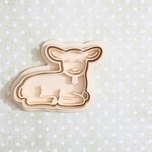 May include: A beige lamb-shaped cookie cutter with a detailed outline. The lamb is in a seated position with its legs tucked beneath it. The cutter has a light brown border and is set against a light green background with white polka dots.