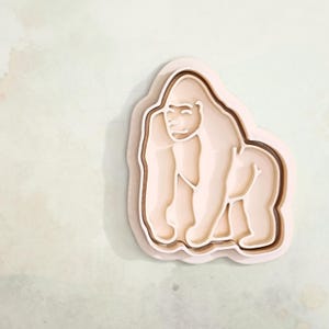 Könnte beinhalten: Ein geschichtetes, hellbraunes Objekt in Form eines Gorillas mit einem lächelnden Gesicht und detaillierten Körpermerkmalen. Der Gorilla ist vor einem hellgrünen Hintergrund mit dezenten aquarellartigen Markierungen platziert. Das Design hat einen 3D-Effekt.