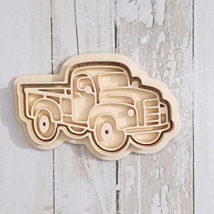 May include: A light beige cookie cutter shaped like a vintage pickup truck. The design features a detailed outline of the truck body, including the cab, cargo bed, wheels, and front grille, with a raised edge for cutting dough shapes.