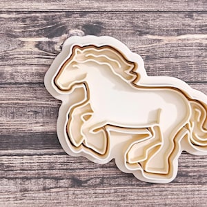 May include: A multi-layered horse-shaped cookie cutter in white, cream, and brown. The design depicts a running horse with a detailed mane and tail. The cutter is made of plastic and is suitable for baking or crafting.