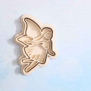 May include: A layered wooden fairy cutout in shades of beige and brown. The fairy has large wings and is depicted in a flying pose. The cutout is on a light blue background with faint script writing.