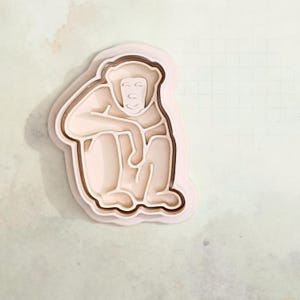 Könnte beinhalten: Hellbeige Affen-Ausstecher. Der Ausstecher hat ein mehrschichtiges Design mit einer detaillierten Kontur eines sitzenden Affen. Der Affe ist mit den Armen auf den Knien dargestellt. Der Ausstecher ist aus Kunststoff.