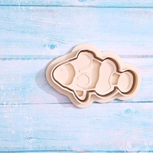 May include: A white plastic cookie cutter shaped like a clownfish. The cutter has a double layer design, with a smaller inner layer for cutting out the fish shape and a larger outer layer for creating a raised edge around the fish.