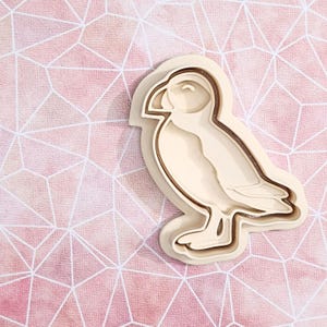 Könnte beinhalten: Hellbeige puffinförmiger Ausstecher. Der Ausstecher hat eine detaillierte Kontur des Vogels, einschließlich Schnabel, Flügel und Füße. Der Ausstecher besteht aus einem hellfarbigen Material und befindet sich vor einem rosa geometrischen Hintergrund.