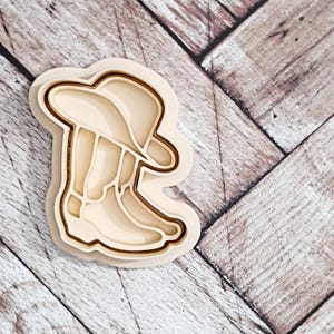 May include: A wooden cookie cutter in the shape of a cowboy boot and hat. The cutter is made of natural wood and has a smooth finish.