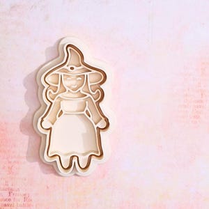 Può includere: Un tagliapasta in plastica bianca a forma di strega con cappello e vestito. Il tagliapasta è su uno sfondo rosa.