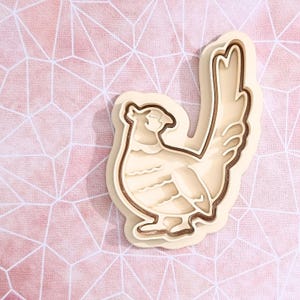 May include: A cream-colored cookie cutter shaped like a pheasant bird. The cutter features a raised outline and internal details that define the bird's head, body, wings, and long tail feathers. The design is molded with a smooth finish.