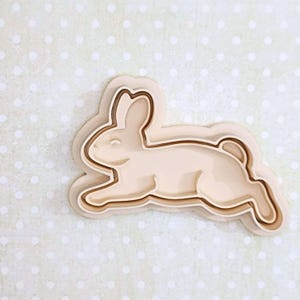 Może przedstawiać: Jasnobeżowa wykrawaczka do ciastek w kształcie królika z trójwarstwowym wzorem. Królik przedstawiony jest w pozycji biegu, z uniesionymi uszami. Wykrawaczka do ciastek znajduje się na jasnym zielonym tle z białymi kropkami.