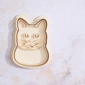 Op de afbeelding: Een houten koekjesvormpje in de vorm van een kattenkop. De vormpje heeft een gedetailleerde omtrek van het kattengezicht, inclusief ogen, neus en snorharen.