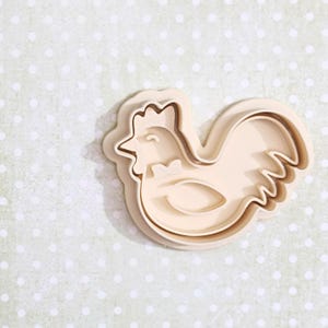 Puede incluir: Cortador de galletas de plástico beige con forma de gallo. El cortador tiene un contorno detallado del cuerpo del gallo, incluyendo su cresta, pico y plumas de la cola. El fondo es de color verde claro con lunares blancos.