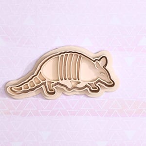 May include: A layered, light brown armadillo-shaped cookie cutter. The cutter has a detailed design with segmented plates, a pointed snout, and small ears. The background is a light pink paper with a repeating triangle pattern.