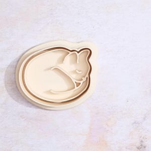 May include: A white plastic cookie cutter shaped like a sleeping cat. The cutter has a double layer design.