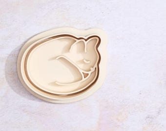 Puede incluir: Un cortador de galletas de plástico blanco con forma de gato durmiendo. El cortador tiene un diseño de doble capa.