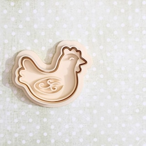 May include: A beige chicken-shaped cookie cutter with a detailed outline and decorative wing design. The cutter is set against a light green background with white polka dots, ideal for baking themed treats.