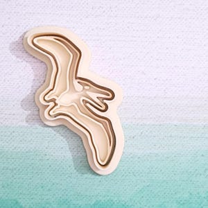 Peut inclure: Un emporte-pièce en bois en forme de ptérodactyle, un dinosaure volant au long cou et aux ailes. L'emporte-pièce est composé de trois tailles différentes, créant un design détaillé.