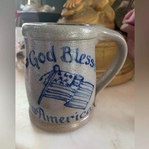 Pode incluir: Uma caneca de grés cinzenta com um acabamento texturizado e salpicado, com uma pega robusta. A parte frontal apresenta o texto pintado à mão em azul God Bless America, acompanhado por uma ilustração azul de uma bandeira americana.