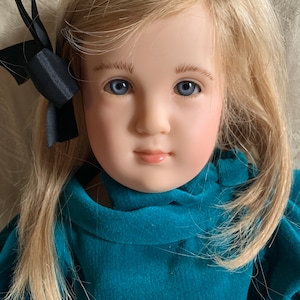 Puede incluir: Una muñeca de porcelana con cabello rubio y ojos azules, vestida con un vestido de terciopelo turquesa con un lazo negro en el pelo.