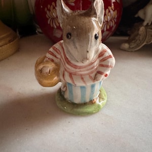 May include: A ceramic mouse figurine wearing a striped top and apron, carrying a basket. The mouse is brown and white, with a green base. A red and gold decorative vase is in the background.