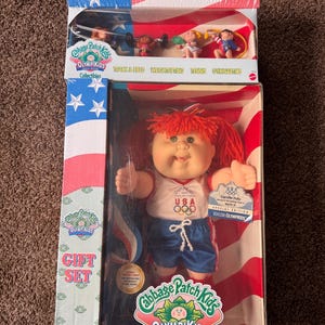 May include: Cabbage Patch Kids OlympiKids gift set. The box features a doll in a white shirt with "USA" and Olympic rings, blue shorts, and red hair. The box also includes miniature sports figures.