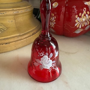 Puede incluir: Una campana de cristal de color rojo intenso con un diseño floral blanco pintado a mano en el exterior. La campana tiene un acabado liso y translúcido y un mango fino y alargado, que muestra un delicado motivo de rosa con hojas.