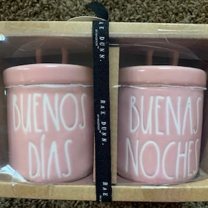 Może przedstawiać: Zestaw dwóch różowych ceramicznych słoików z pokrywkami, każdy z białym tekstem. Jeden głosi "BUENOS DÍAS", a drugi "BUENAS NOCHES". Czarna wstążka z białym tekstem "RAE DUNN" znajduje się między słoikami.