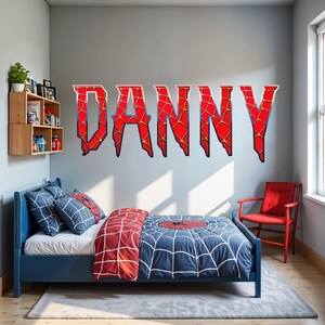 Pode incluir: Um adesivo de parede com o nome DANNY em letras grandes, vermelhas e irregulares com um padrão de teia de aranha preta. As letras estão aplicadas numa parede cinzenta clara acima de uma estrutura de cama azul com roupa de cama de teia.