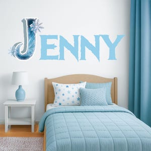 Pode incluir: Um decalque de parede azul claro com o nome "JENNY" em um quarto. A letra "J" é decorada com um floco de neve e um efeito aquarela azul. A cama tem roupa de cama azul e travesseiros com estampa de estrelas.