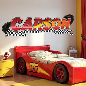 Peut inclure: Un autocollant mural affichant le nom CARSON en lettres rouges et orange avec un éclair jaune, sur un fond de drapeau à damier noir et blanc, surmonté d'un graphique représentant une route courbe au-dessus du texte.