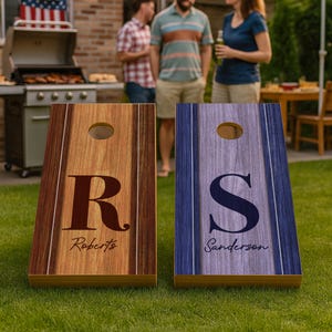 Pode incluir: Duas tábuas de cornhole de madeira com designs personalizados. Uma tábua é castanha com a letra "R" e o nome "Roberts". A outra é azul com a letra "S" e o nome "Sanderson". Ambas as tábuas têm um buraco na parte superior.