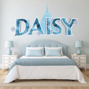 May include: A bedroom scene featuring a bed with a light blue headboard, white bedding, and a blue bed runner. Above the bed, the name "DAISY" is displayed in large, icy blue letters with snowflake accents and a castle silhouette.