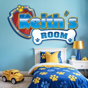May include: A child's bedroom with a blue wall featuring a wall decal that reads "Keith's ROOM" in blue and white. The bedding is blue with yellow paw prints. A yellow toy truck sits on the floor.