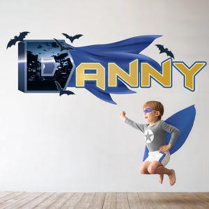 Pode incluir: Um decalque de parede com o nome "DANNY" em letras douradas e um design de capa azul. O "D" tem um fundo de paisagem urbana e silhuetas de morcegos. Uma criança fantasiada de super-herói saltando.