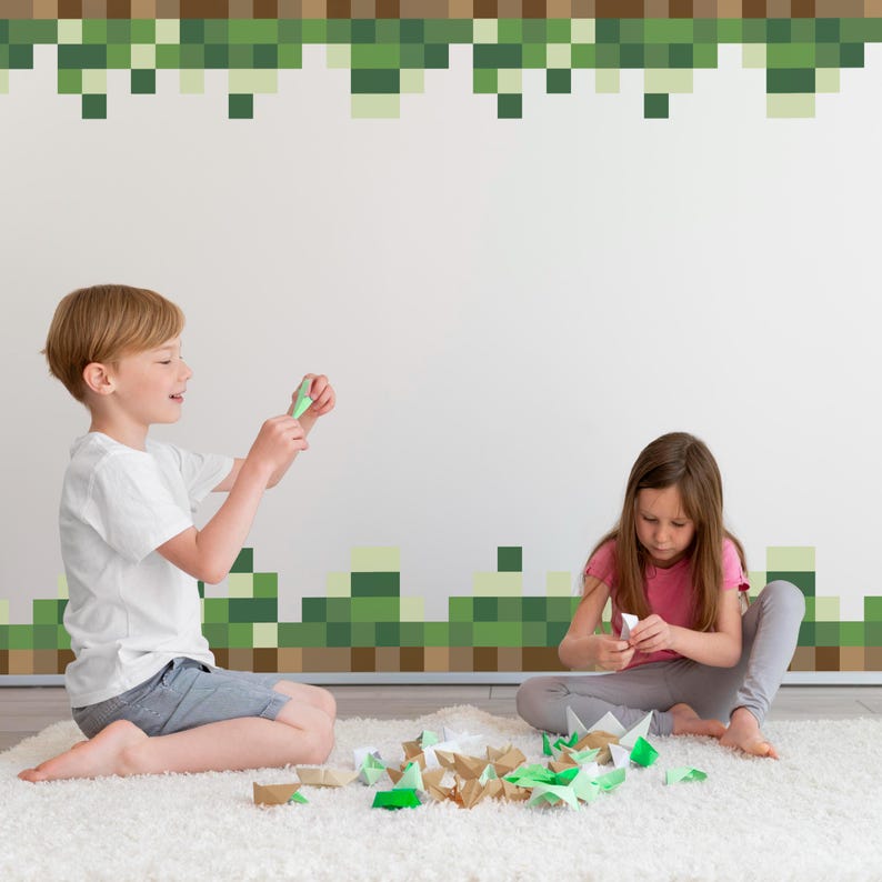 May include: Two children are seated on a white rug, surrounded by folded paper shapes. The boy is holding a green paper shape, while the girl is working on another. The wall behind them has a pixelated green and brown design.