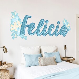 Pode incluir: Um decalque de parede com o nome "Felicia" em uma fonte cursiva azul claro brilhante. Acentos florais em azul claro e creme circundam o nome. O decalque está acima de uma cama com travesseiros azuis e um edredom branco.