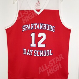 Op de afbeelding: Rood basketbaltenue met witte afwerking en de tekst "Spartanburg 12 Day School" in blauwe letters. Het tenue heeft een witte V-hals en een klein rood-wit logo op de voorkant.