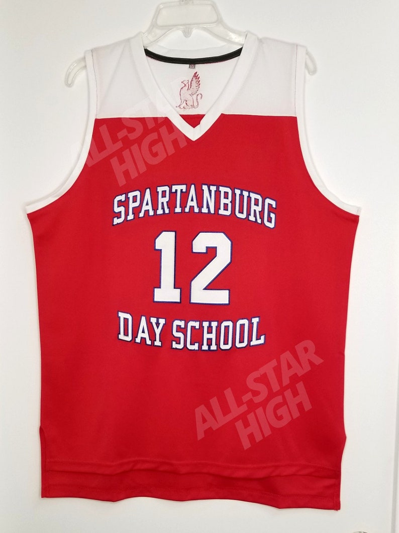 Op de afbeelding: Rood basketbaltenue met witte afwerking en de tekst "Spartanburg 12 Day School" in blauwe letters. Het tenue heeft een witte V-hals en een klein rood-wit logo op de voorkant.