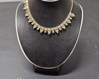 May include: A gold-toned jewelry set, including a necklace, bracelet, and earrings, displayed on a black mannequin. The necklace and bracelet feature a repeating pattern of clear, sparkling stones. The earrings are a curved design with similar stones.