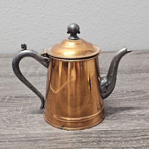 May include: A small, antique copper teapot with a silver-colored handle, spout, and lid. The teapot has a rounded body and a small, decorative knob on the lid. The copper has a warm, reflective finish, and the teapot sits on a wooden surface.