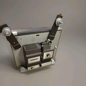 May include: A vintage gray and black film projector featuring two upward-extending reel arms with silver spindles. The central housing includes a black vented cover, a silver control panel with a black knob, and a label reading EUMIG MARK 501.