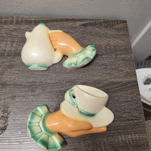 May include: Two ceramic wall hangings, one with a teapot and the other with a teacup and saucer, each held by a hand. The hands are peach-colored, and the teapot, teacup, and saucer are white with green trim.